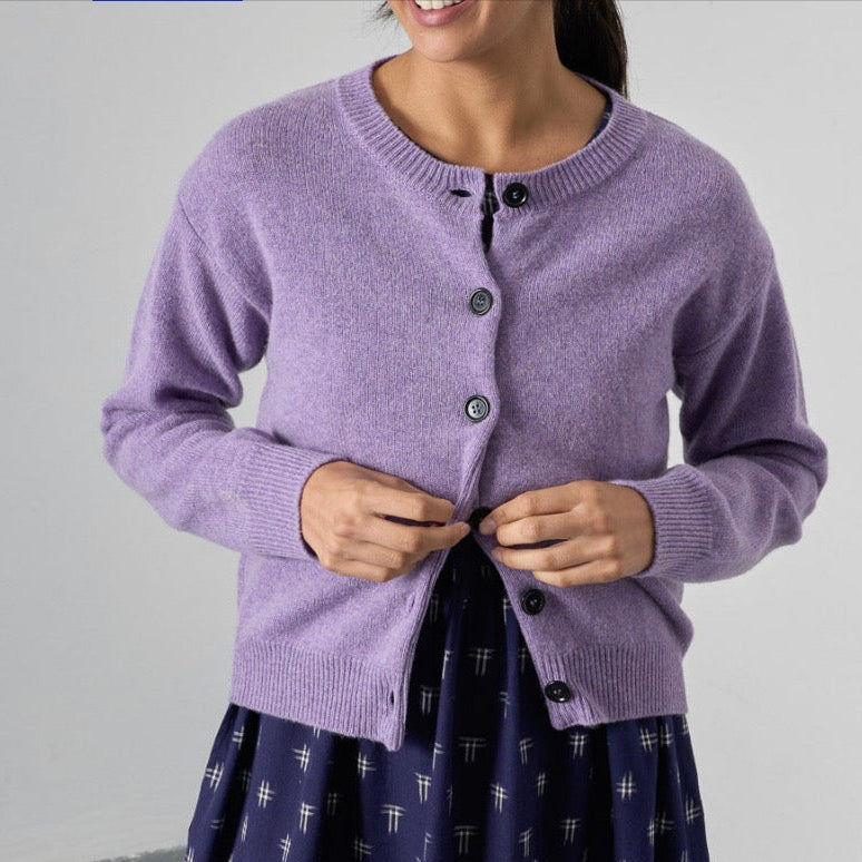 A woman wearing a light purple knit cardigan with dark buttons and a navy patterned skirt, shown from the waist up against a plain light gray wall.