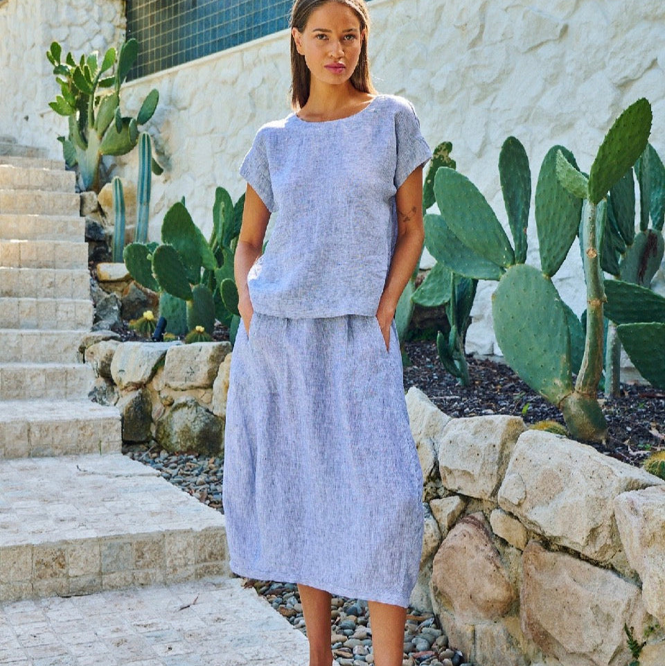 A woman stands outdoors on stone steps beside a stone wall and cactus plants, wearing a light lavender-blue short-sleeve top and matching midi skirt with a subtle textured pattern.