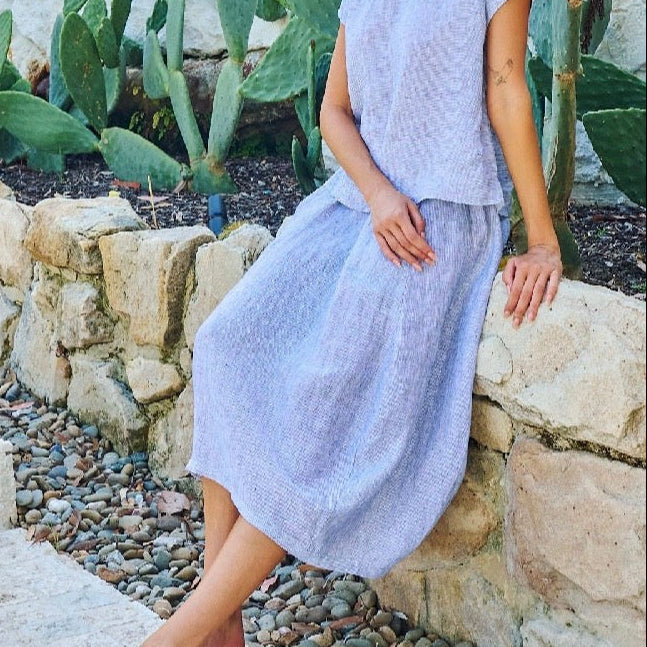 A woman wearing a light lavender textured two-piece set consisting of a short-sleeve top and a midi skirt, leaning against a stone wall in an outdoor garden setting with cacti.