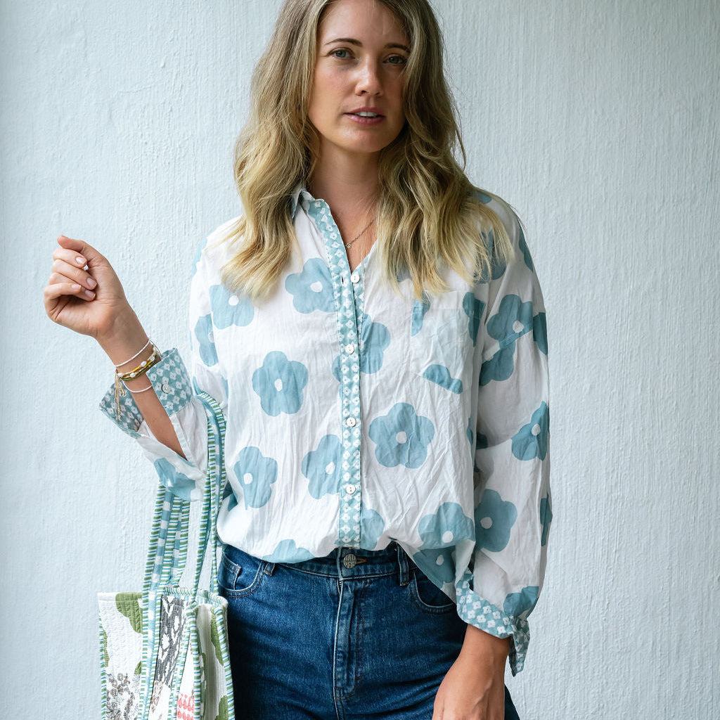 A woman wearing a white long-sleeve button-up blouse with a light blue floral pattern and blue jeans, holding a matching patterned tote bag against a light blue textured wall.