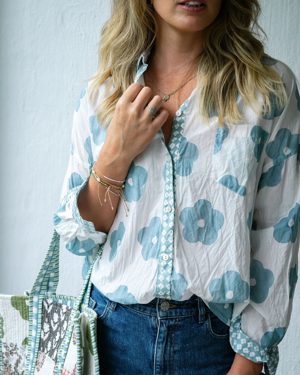 A woman wearing a white long-sleeve button-up blouse with a light blue floral print and matching patterned trim, paired with blue jeans and carrying a patterned tote bag.