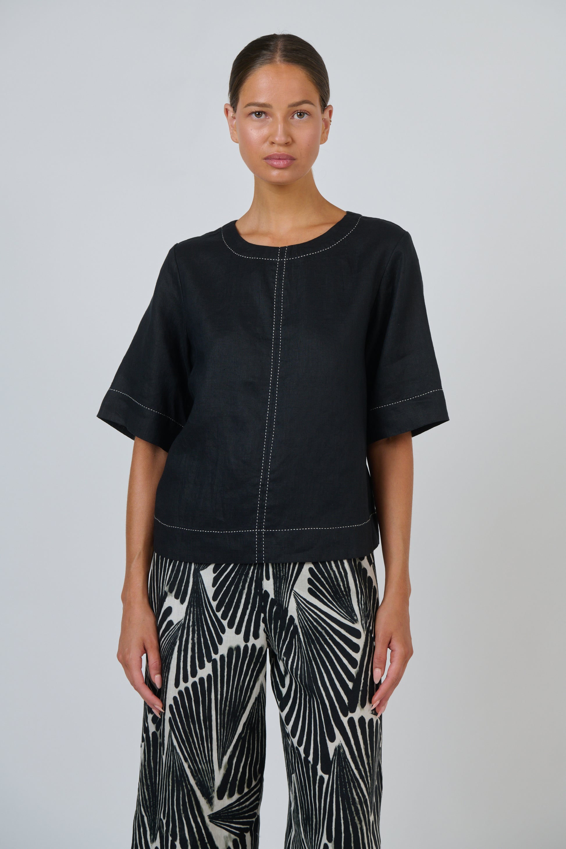 A model wearing a black short-sleeve top with white contrast stitching and black-and-white patterned pants, standing against a light gray studio background.