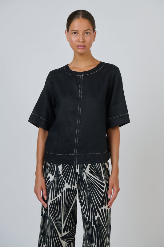 A model wearing a black short-sleeve top with white contrast stitching and black-and-white patterned pants, standing against a light gray studio background.